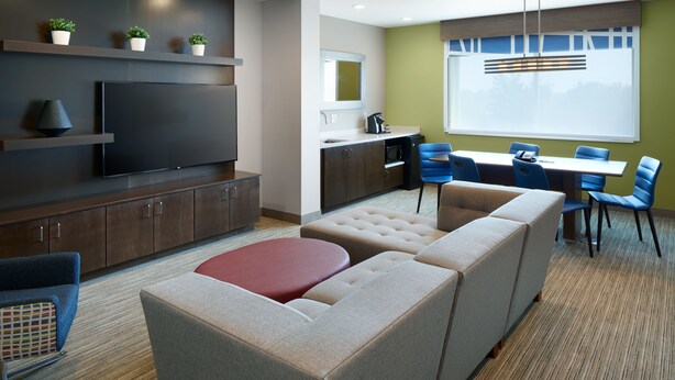 A modern living room with a large sectional sofa, a flat-screen TV, built-in cabinets, a kitchen area, and a dining table with chairs. - Holiday Inn Express & Suites-Cincinnati North - Liberty Way by IHG (West Chester)