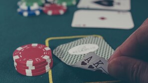 Eine Hand hält eine Spielkarte mit Herzsymbol, ein Stapel rote Pokerchips und eine grüne Spielkarte auf einer grünen Fläche.