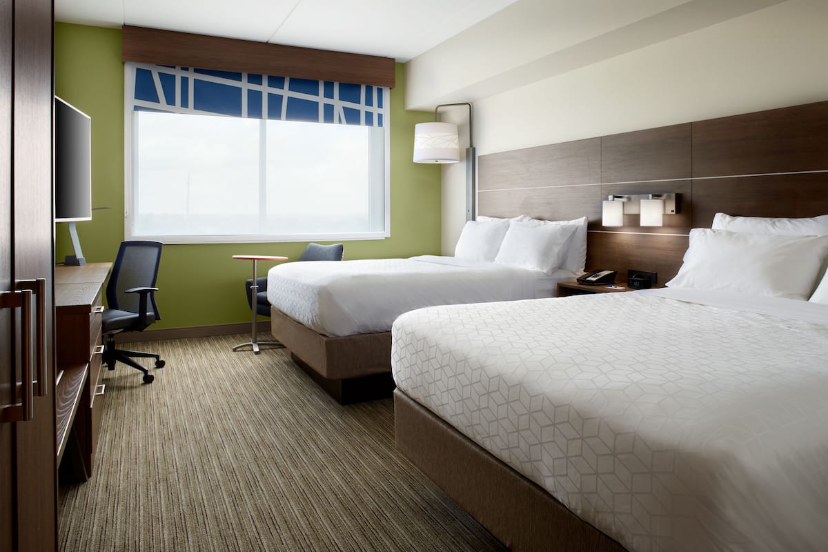 A hotel room with two beds, a desk, a chair, a TV, and a window with blinds.
