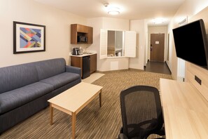 Ein Hotelzimmer mit einer Couch, einem kleinen Tisch, einem Schreibtisch mit Stuhl, einer Mikrowelle, einem Waschbecken und einem Fernseher.