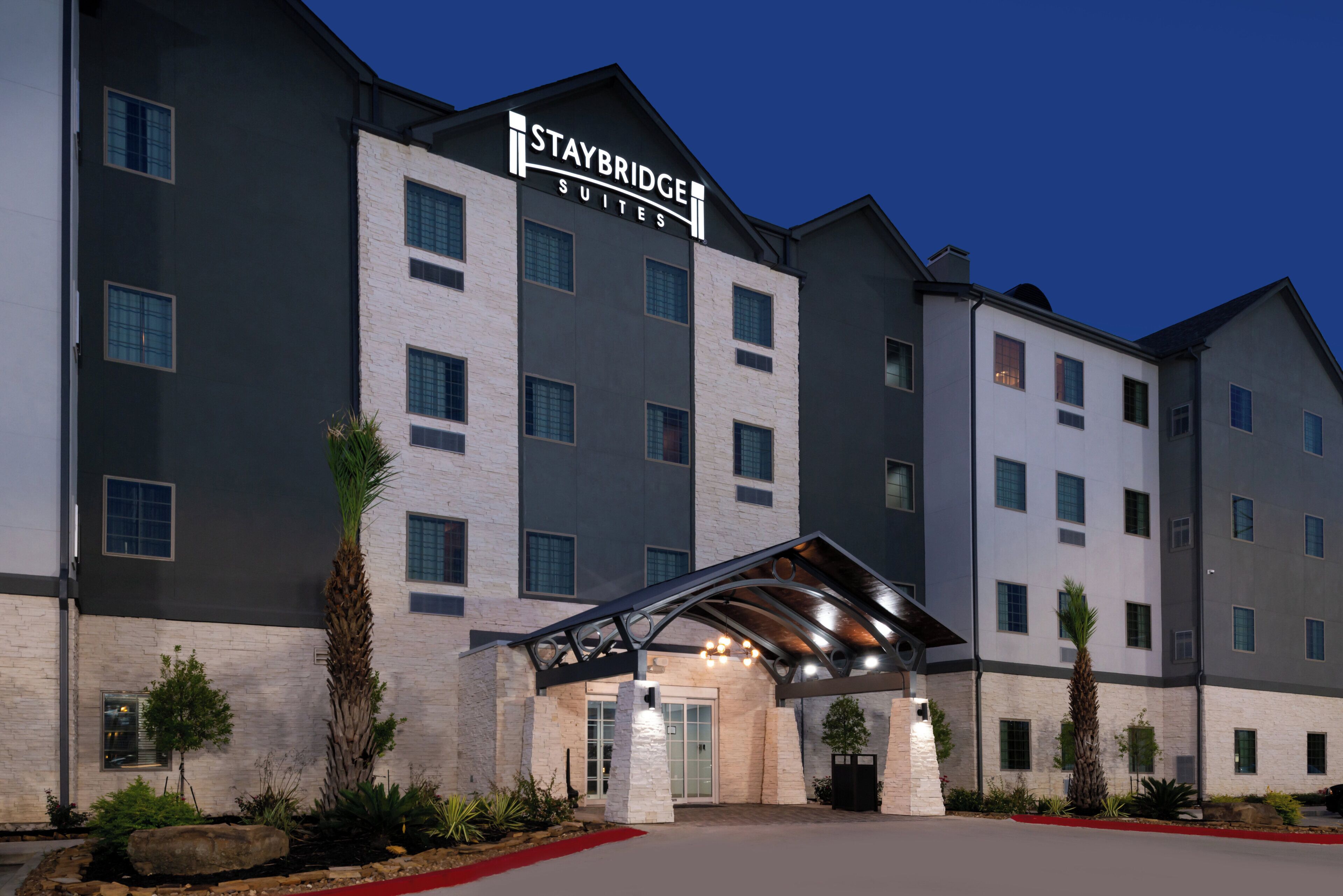 Un hôtel Staybridge Suites au design moderne, avec une entrée imposante dotée d’une allée couverte et un ciel dégagé en arrière-plan.