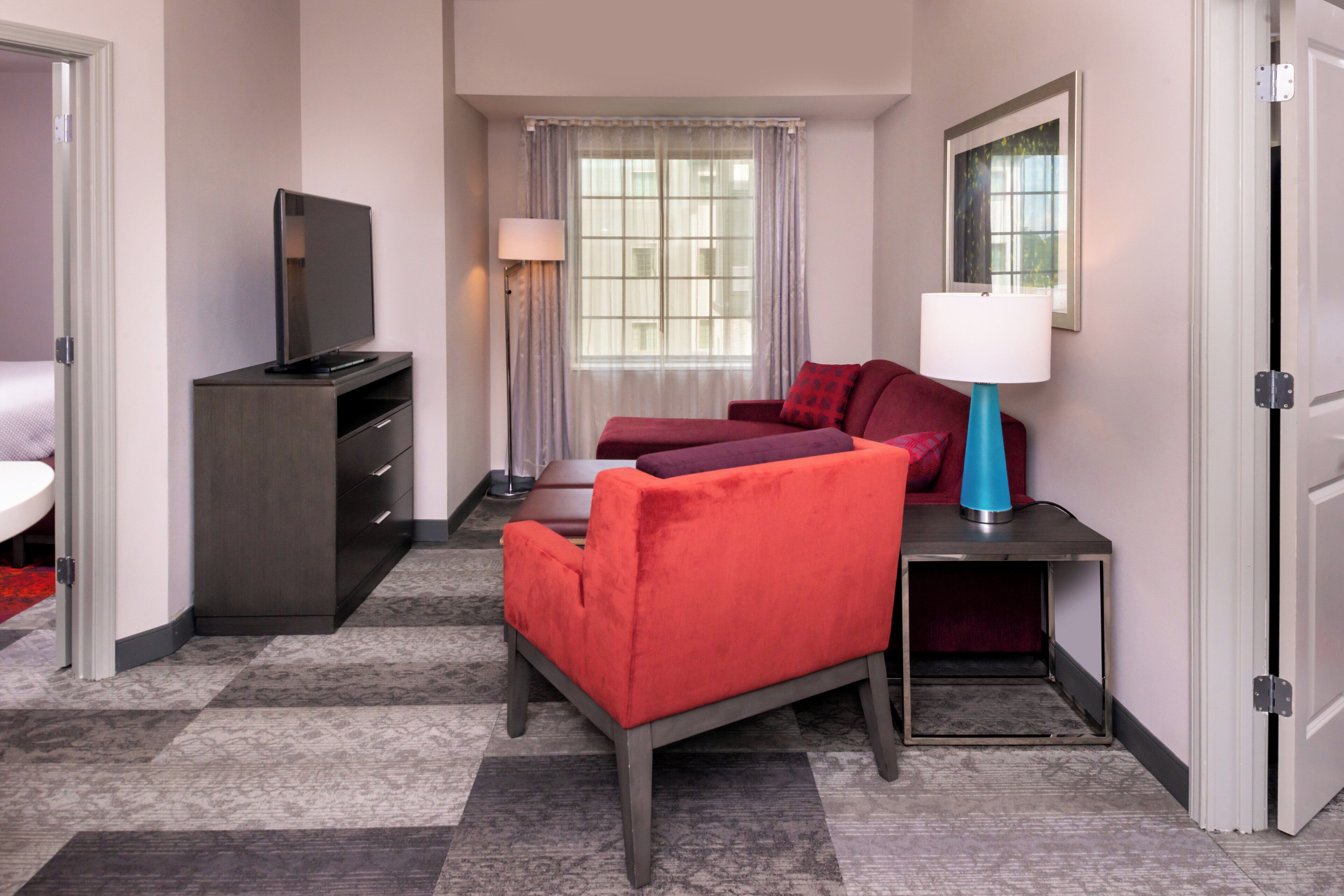 Une chambre d’hôtel avec un lit, un bureau avec une lampe, une télévision et une fenêtre avec des rideaux.