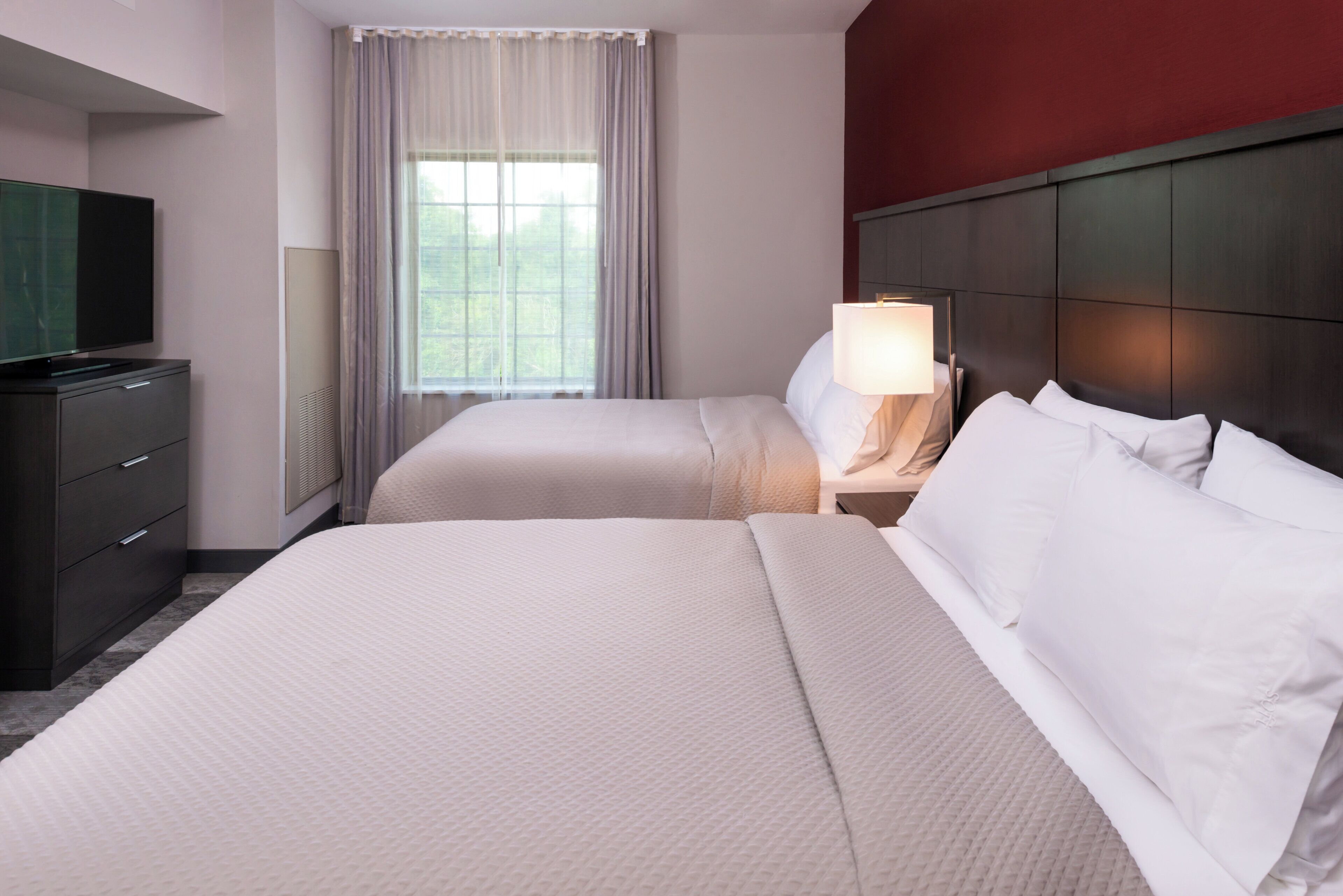 A hotel room with two beds, a television, a nightstand with a lamp, and a window with curtains.