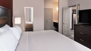 A hotel room with a large bed, a dresser, a television, and a door leading to a kitchen area.