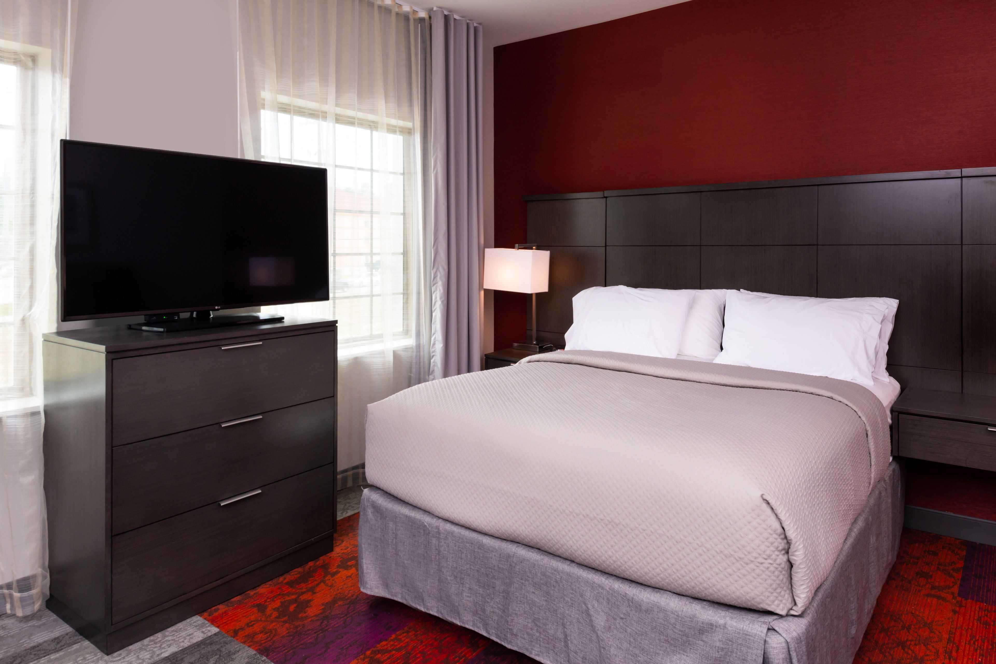 A hotel room with a large flat-screen TV on a dark wooden dresser, a bed with white linens, and a red accent wall.