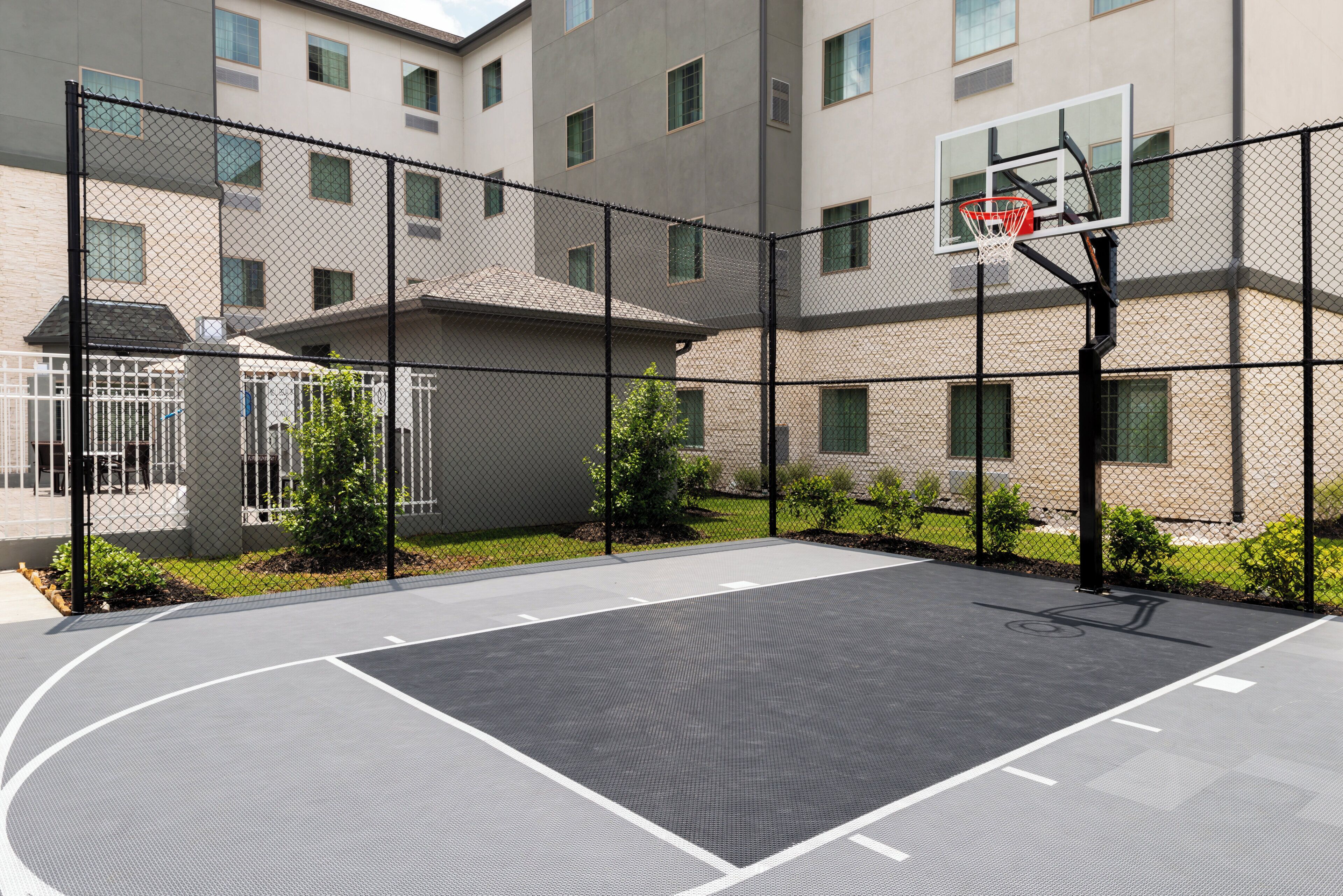 An outdoor basketball court with a chain-link fence, basketball hoop, and a building in the background.