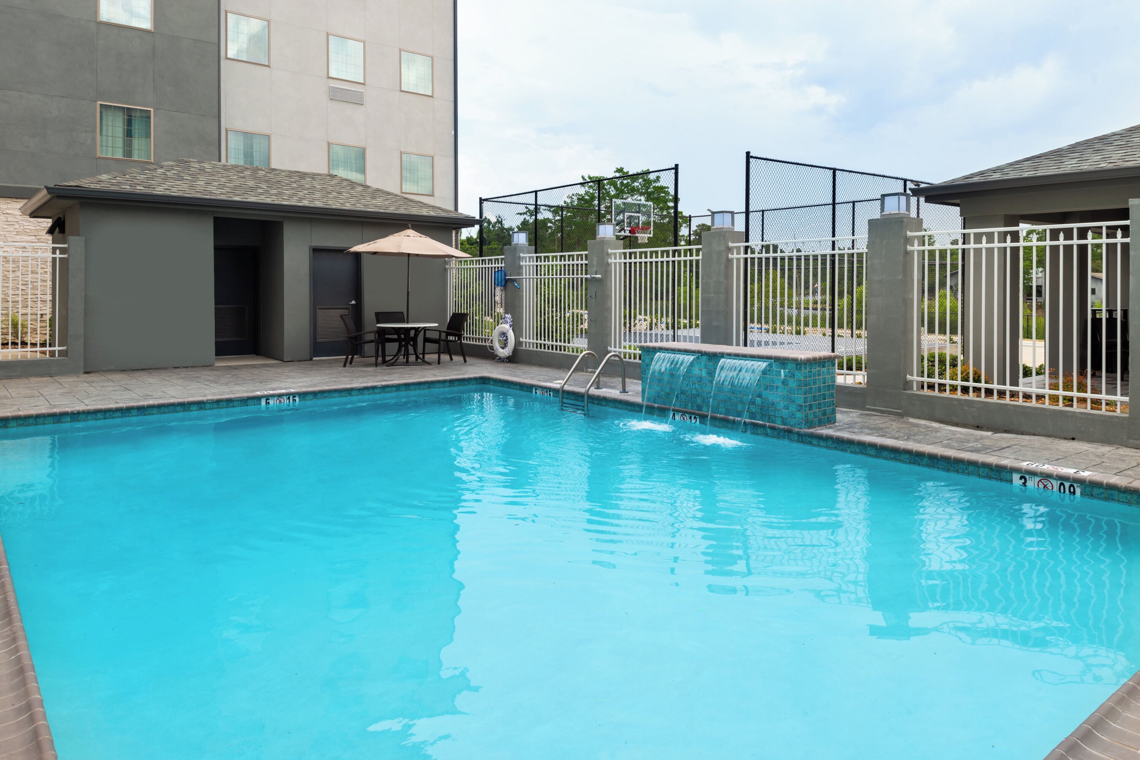 Une piscine avec un plongeoir, un espace de détente avec des chaises et une table, et un panier de basketball.