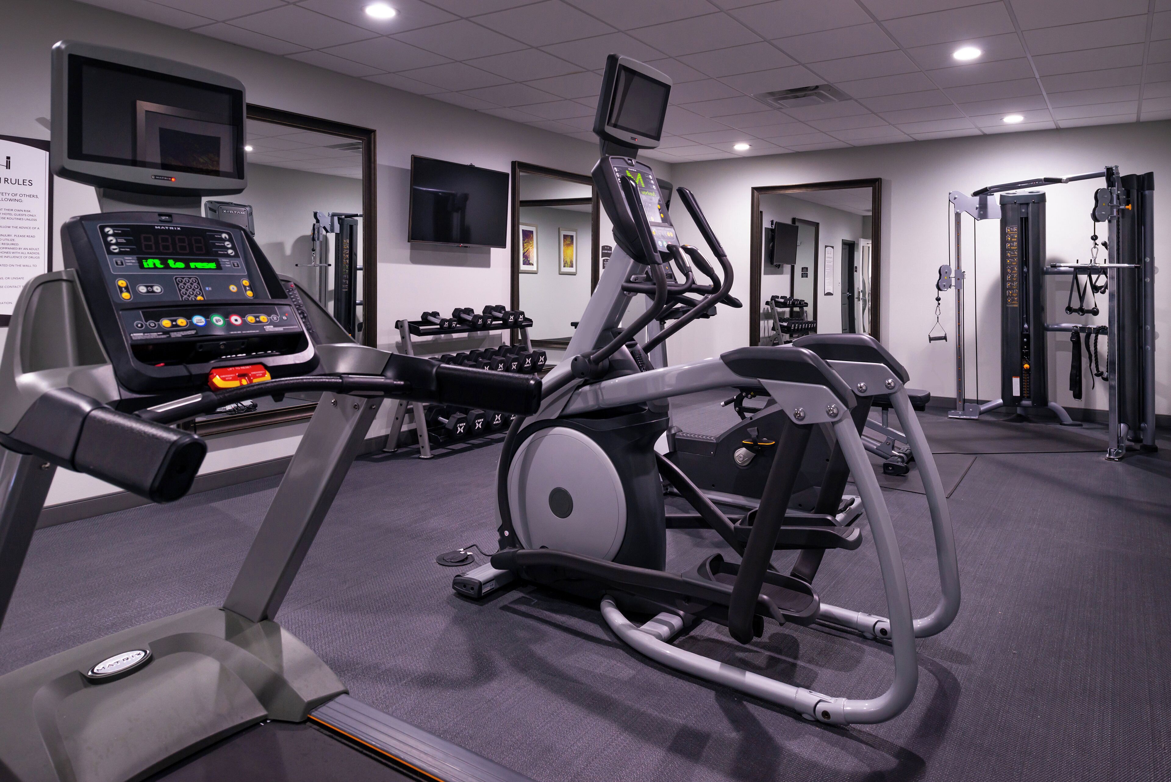 A gym with cardio equipment, dumbbells, and a television screen.
