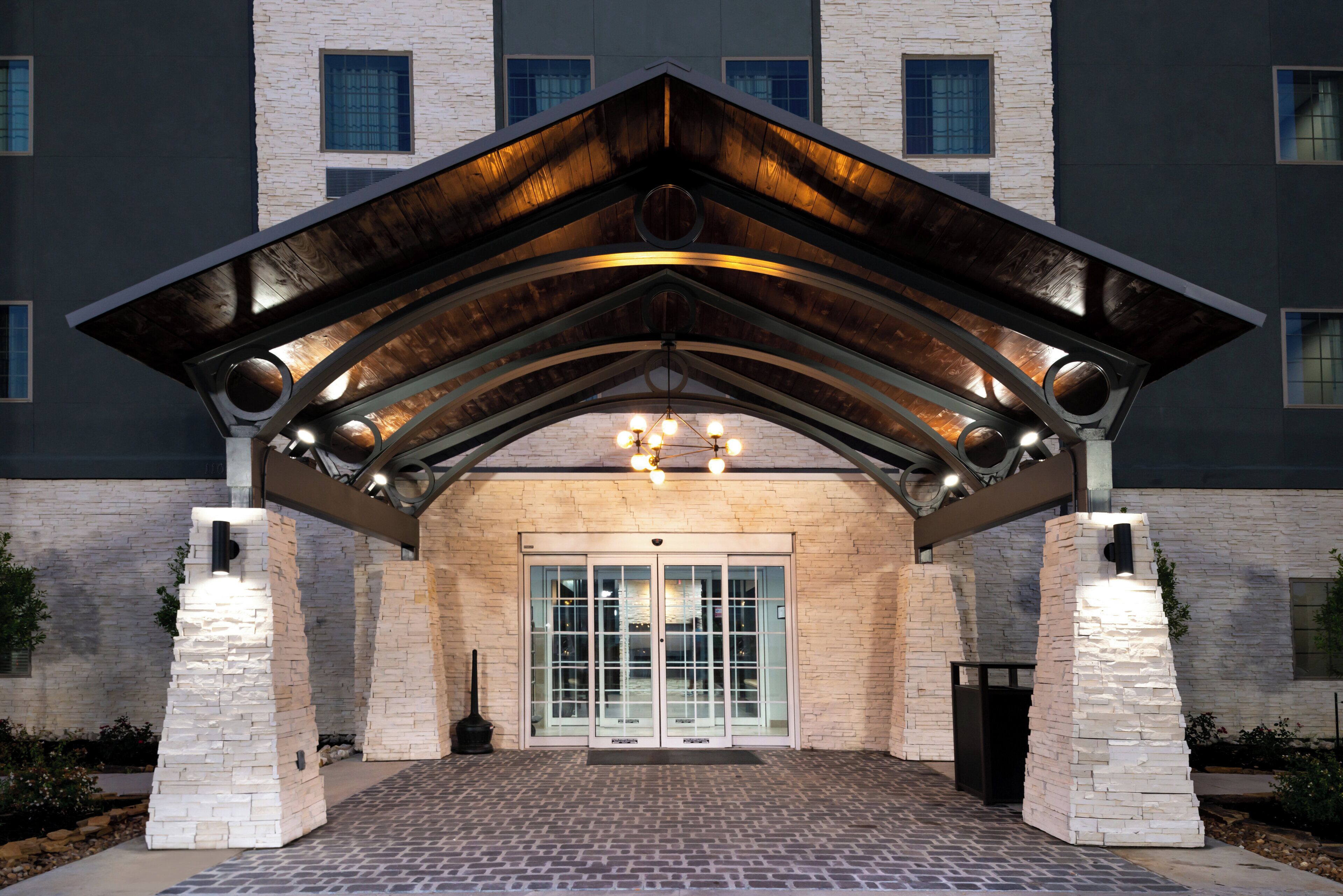 Une entrée d’immeuble avec une façade en pierre, une structure de toit en métal et une porte d’entrée en verre.