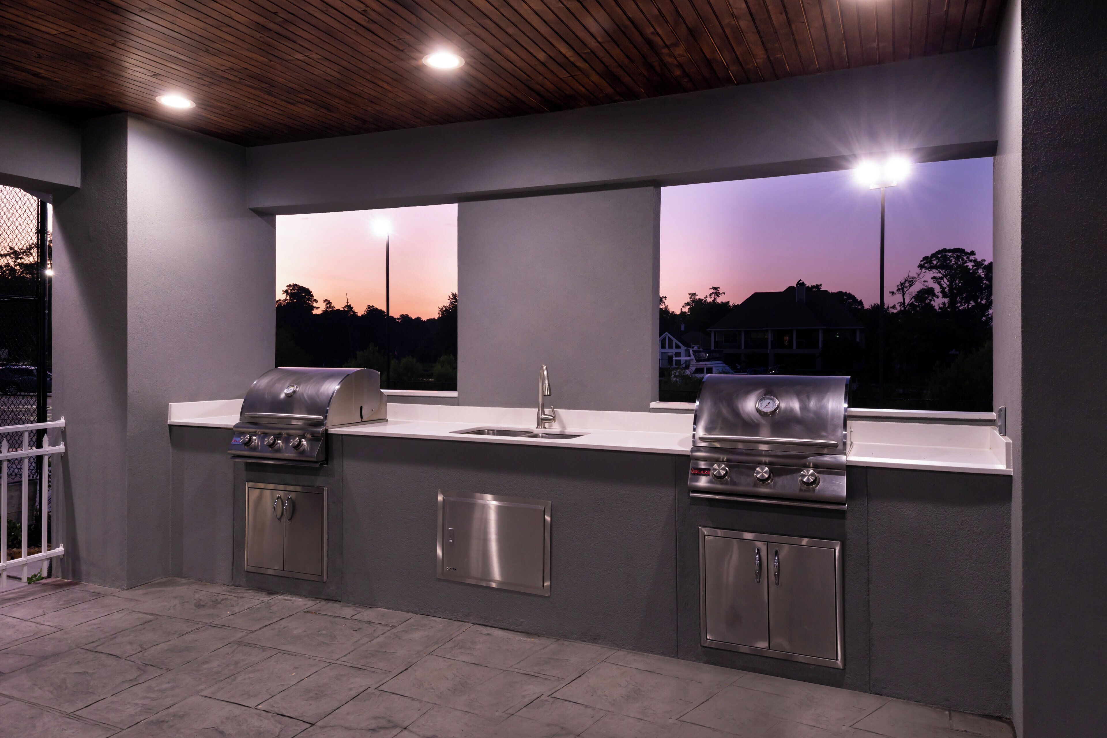 An outdoor kitchen with two gas grills, a sink, and a window overlooking a sunset.