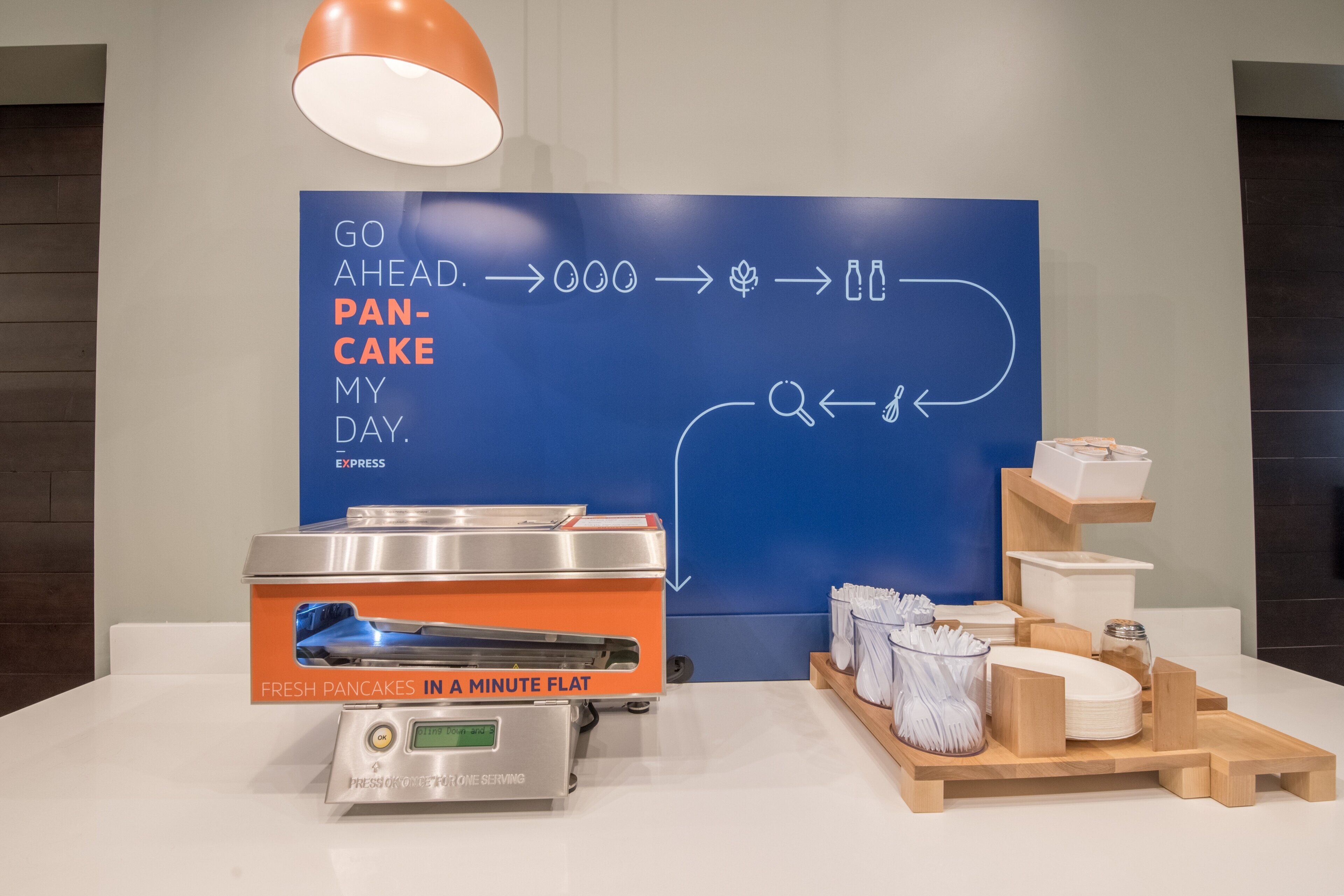 A breakfast setup with a flat-screen TV displaying a breakfast menu, a digital pancake maker, and a tray of breakfast items.