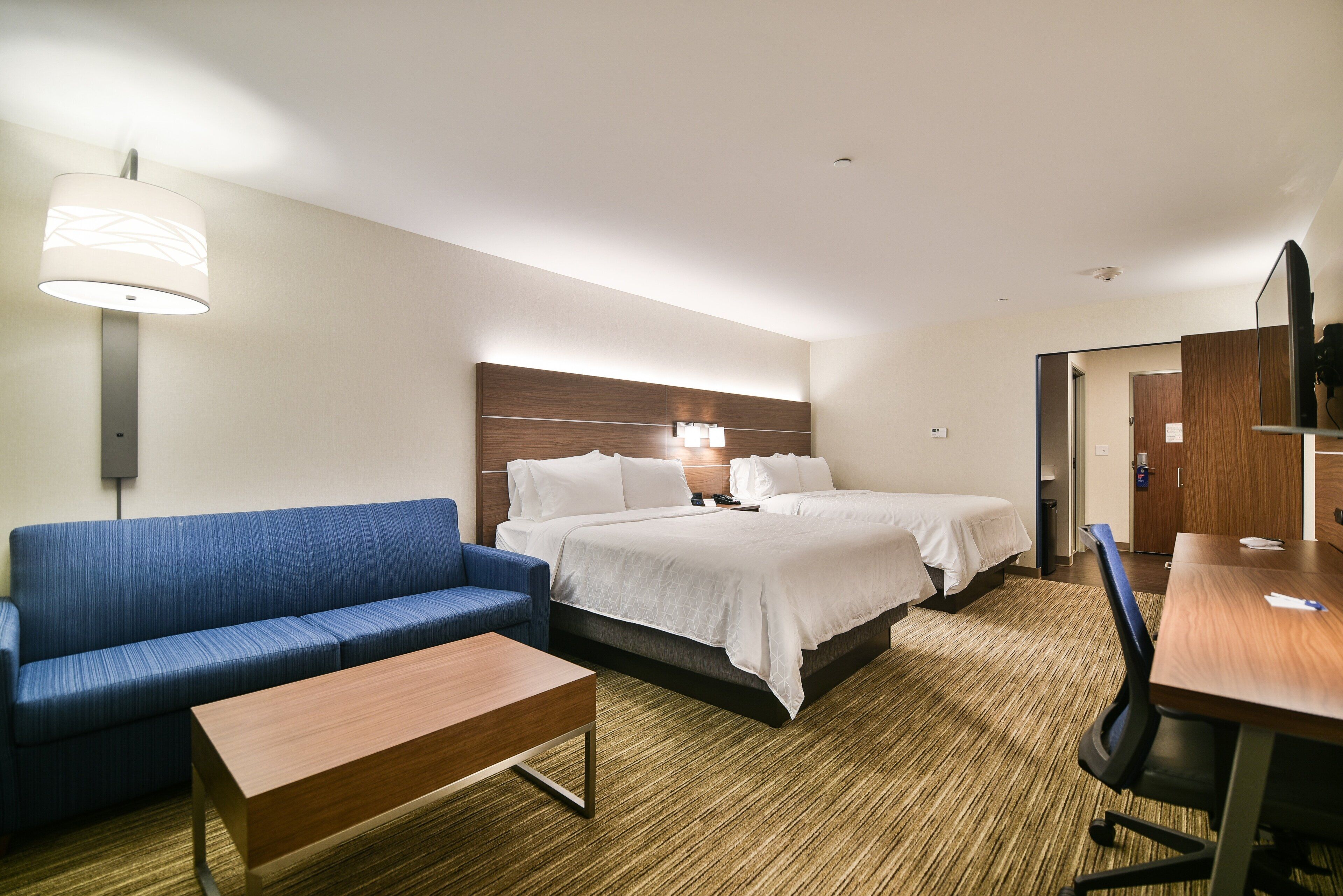 Ein Hotelzimmer mit einem blauen Sofa, einem Holztisch, einem Doppelbett mit weißer Bettwäsche, einem Schreibtisch mit Stuhl und einer Wandlampe.