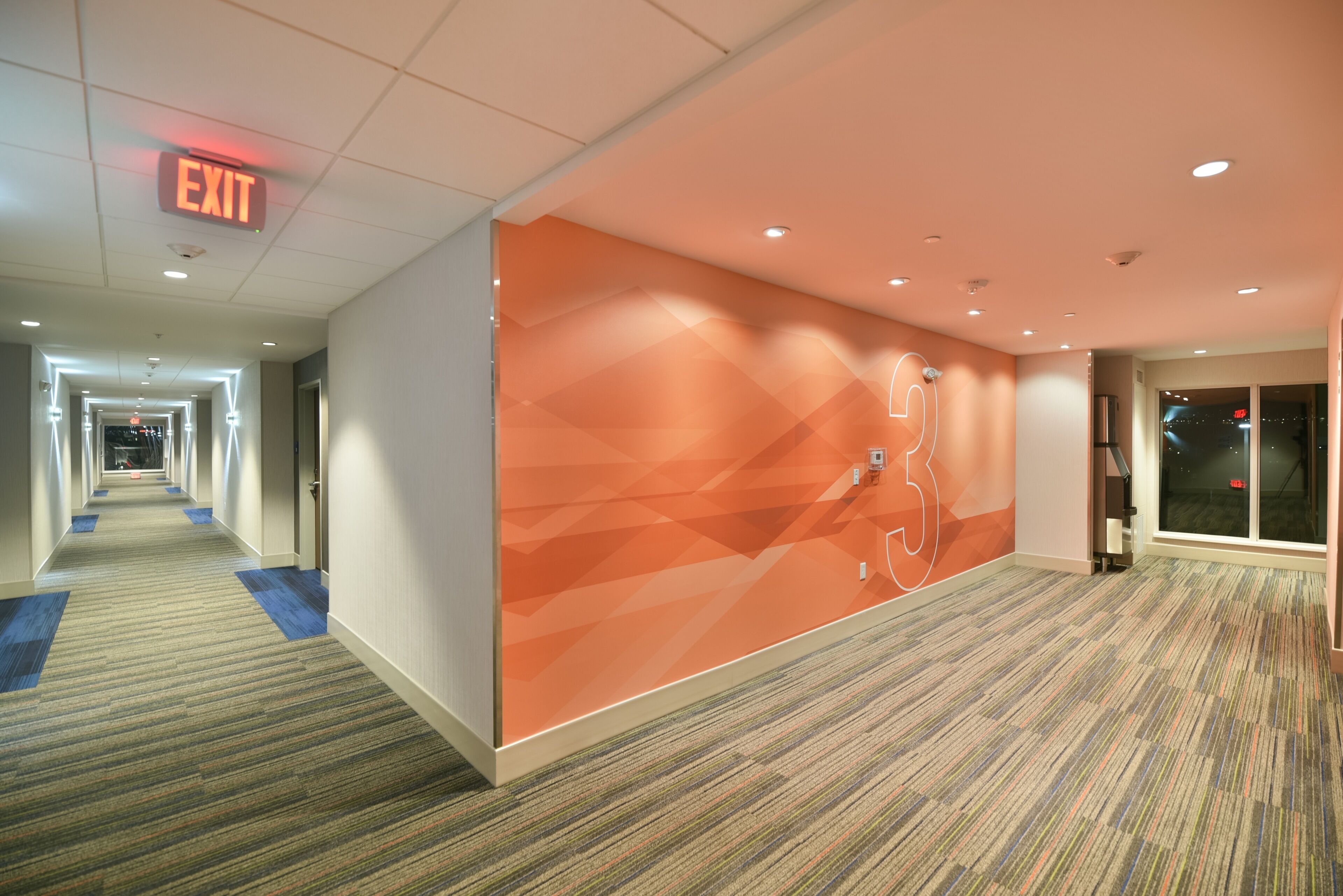A hotel corridor with an exit sign, a geometric wall design, and a carpeted floor.