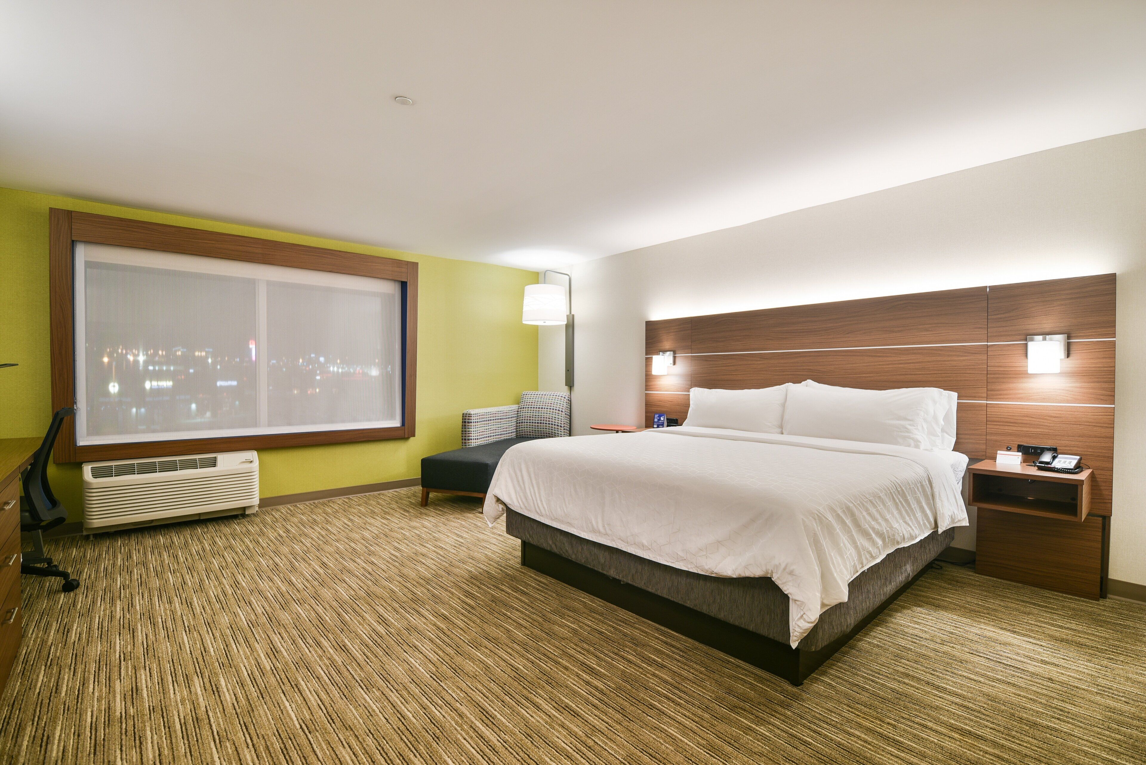 Quarto de hotel com cama grande, mesa de cabeceira, escrivaninha, cadeira, janela com vista para a cidade e luminária na parede.