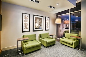 A hotel lobby with three green armchairs, a small table, and framed artwork on the wall.