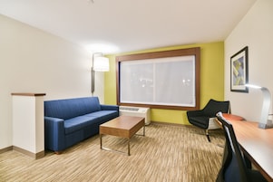 A modern hotel room with a blue sofa, a wooden coffee table, a black chair, and a large window with blinds.