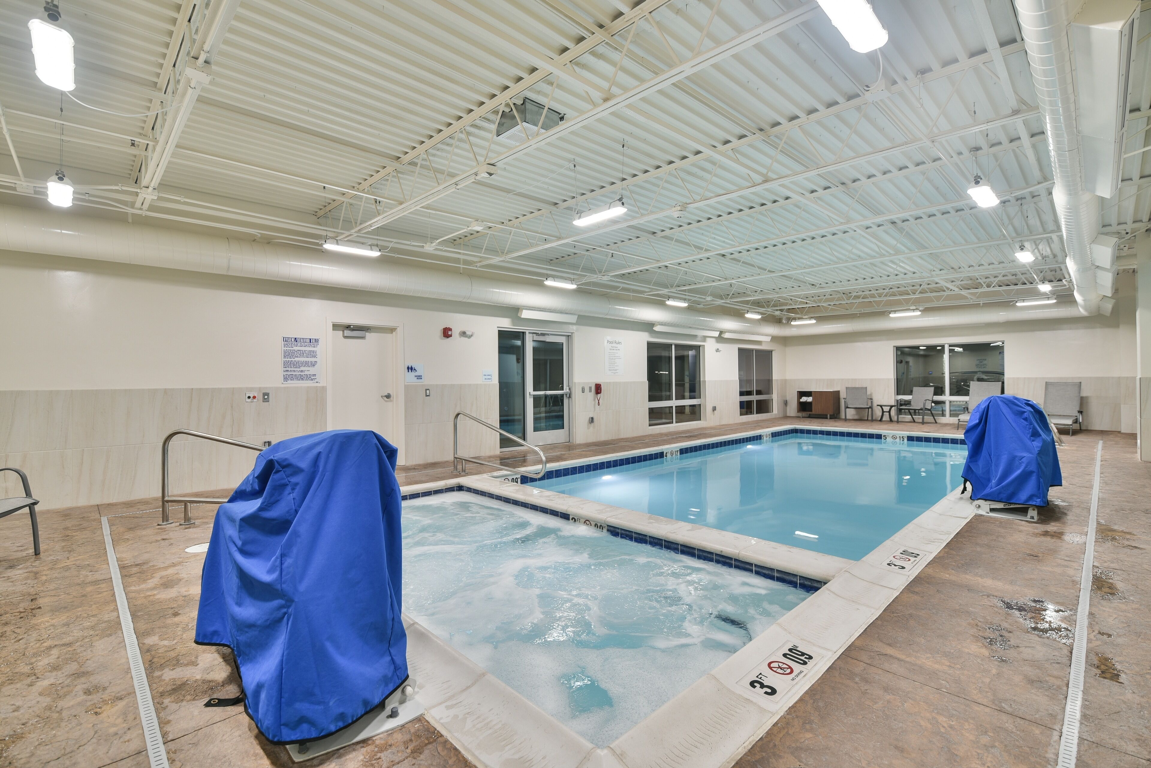 Una piscina interior con agua cristalina, bordes de azulejos y una zona techada con una silla para socorrista.