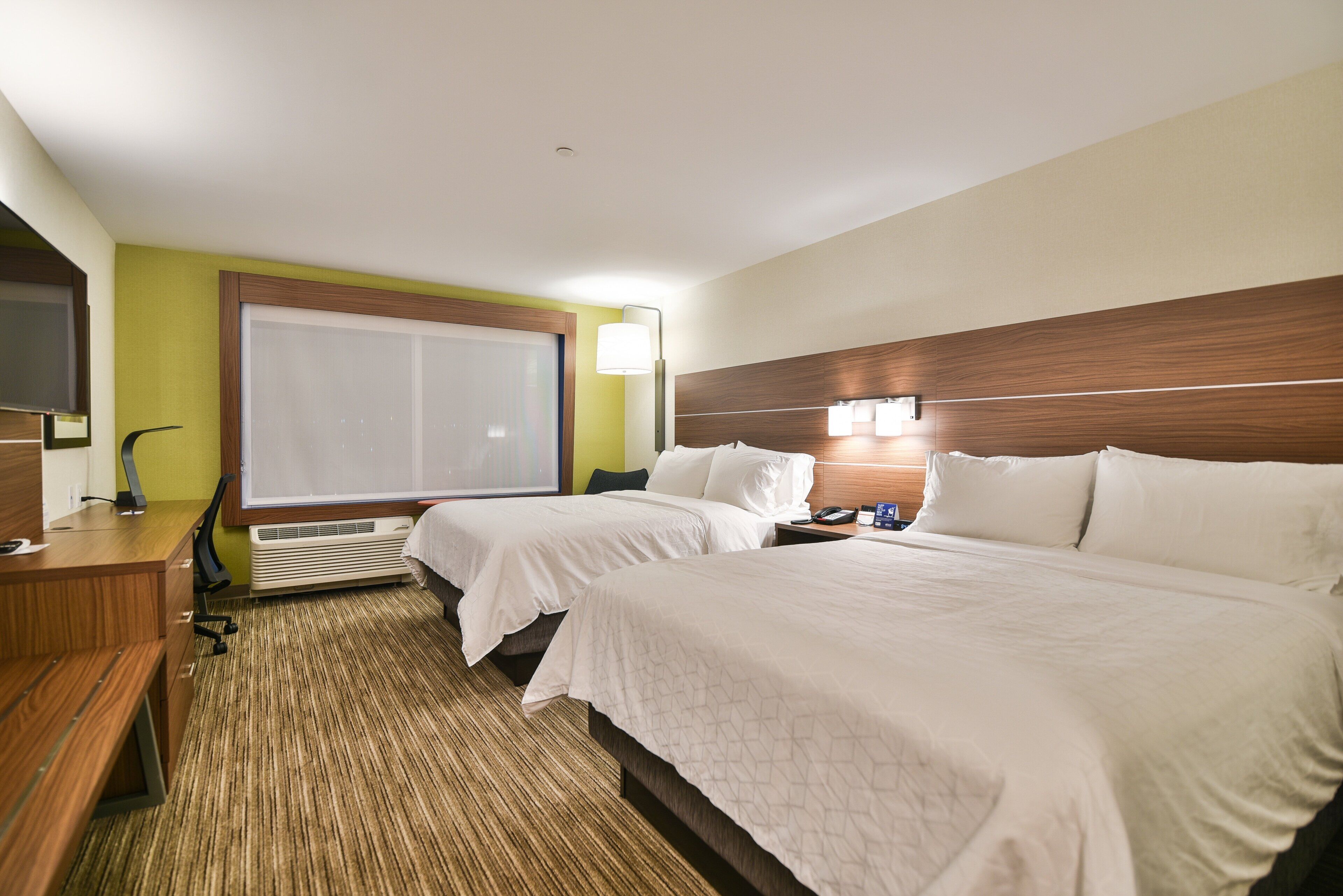 A hotel room with two beds, a desk, a chair, a window with blinds, a standing lamp, and a wall-mounted TV.