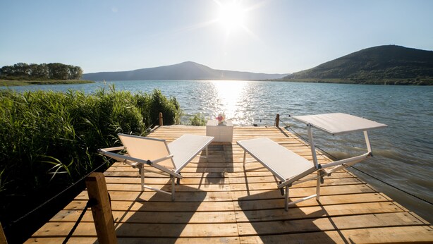 Une terrasse en bois avec deux chaises longues blanches et une petite table, donnant sur un lac paisible avec des montagnes en arrière-plan.