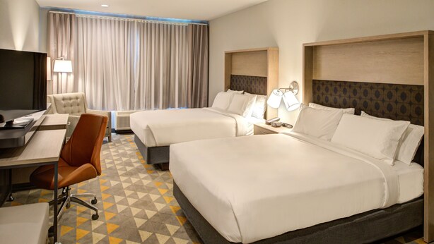 A hotel room with two beds, a desk with a chair, a TV, and a large window with curtains.