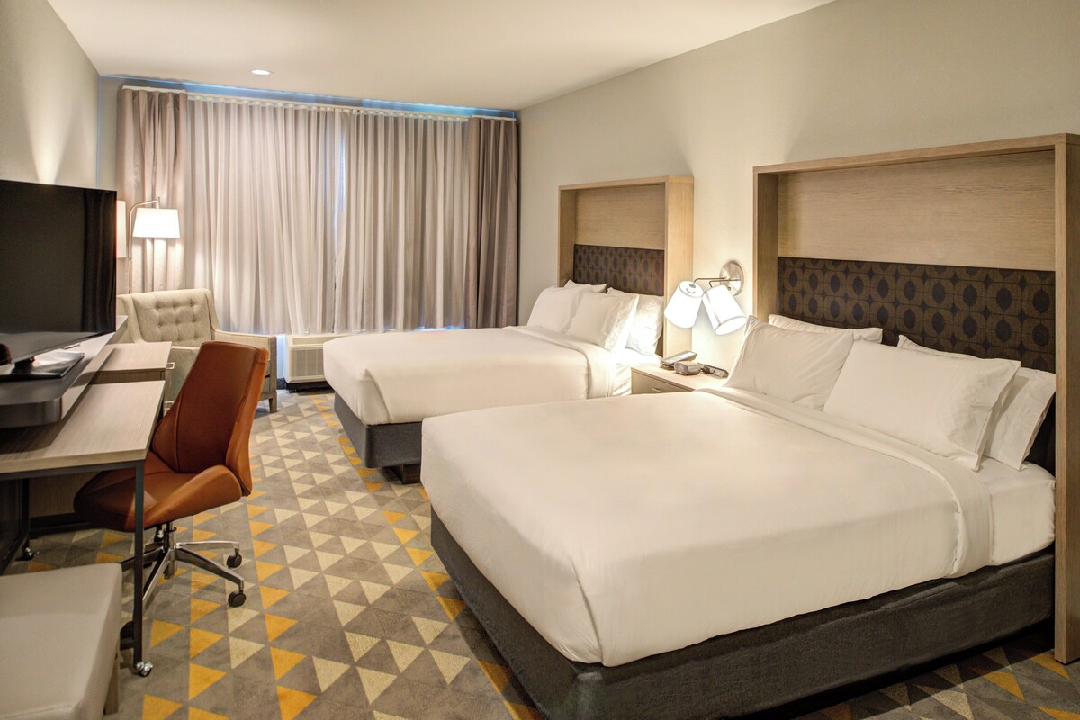 A hotel room with two beds, a desk with a chair, a TV, and a window with curtains.