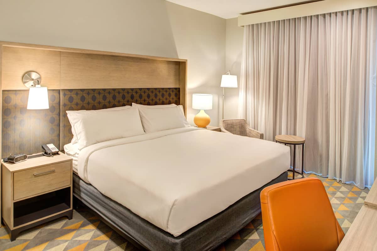 Une chambre d’hôtel avec un grand lit, des tables de chevet, une chaise et une vue sur l’extérieur à travers les rideaux.