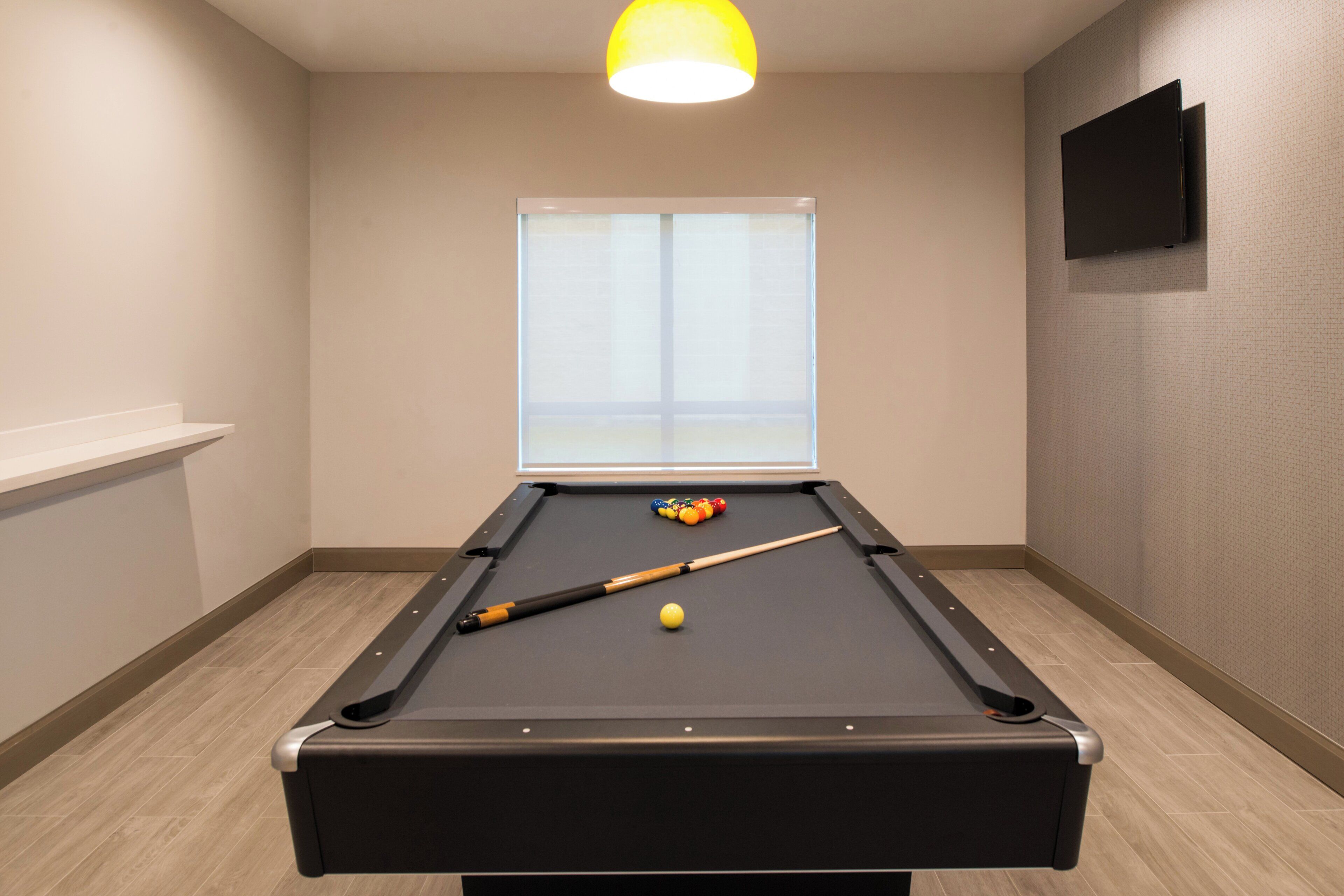 A pool table with a cue stick and billiard balls, a window with blinds, and a flat-screen TV mounted on the wall.