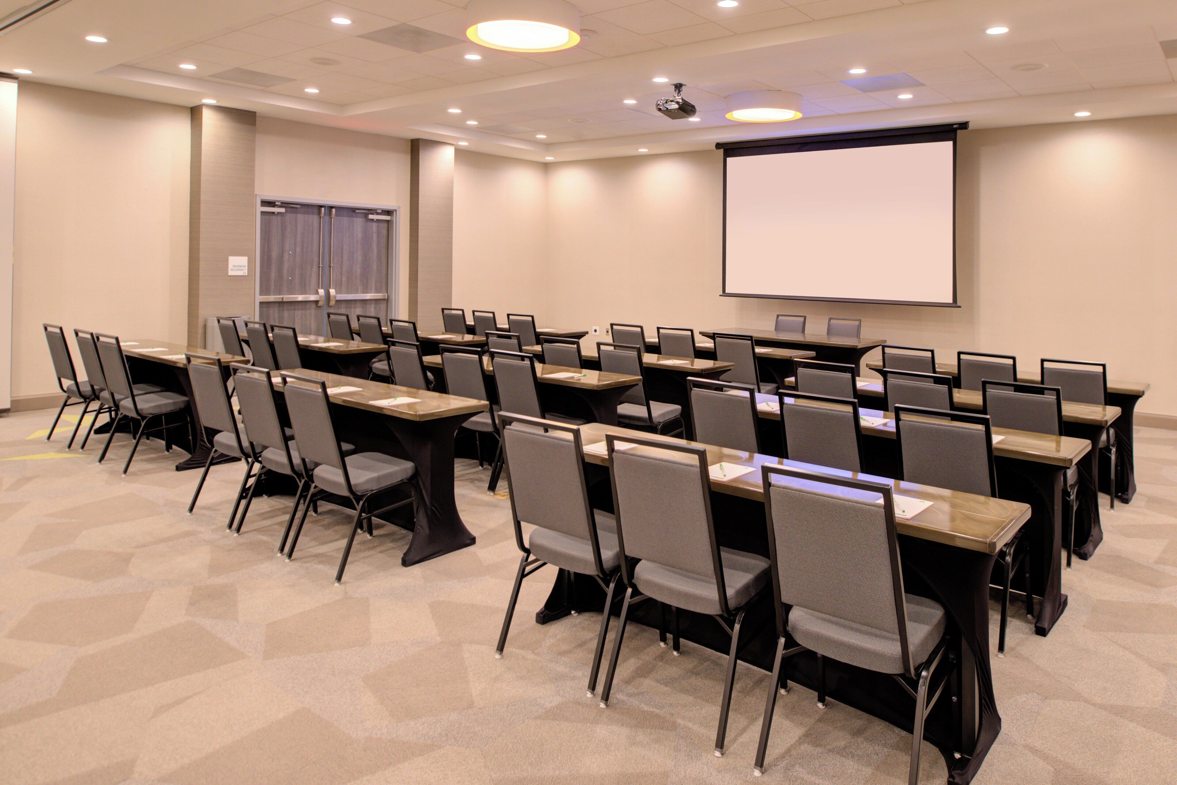 Un salón de conferencias con filas de mesas y sillas, una pantalla de proyector y un proyector instalado en el techo.