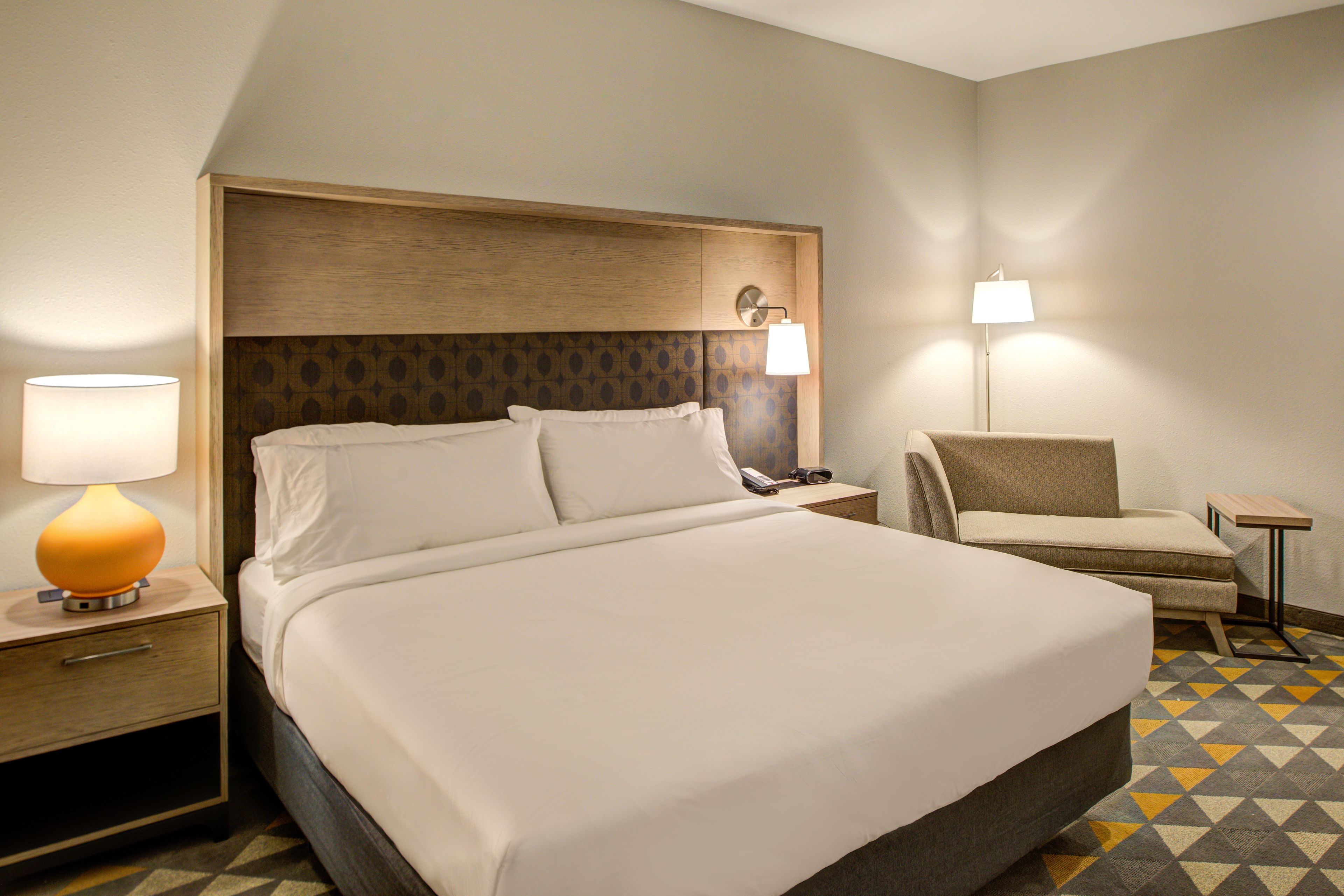 A hotel room with a large bed, a nightstand with a lamp, a chair, and a small table.