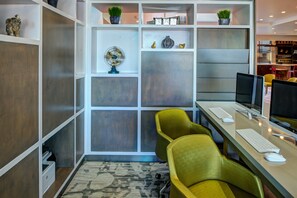 A modern office space with a desk, computer, and two chairs. Shelves with decorative items and a fan are visible.