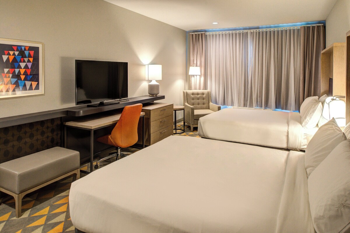 A hotel room with two beds, a desk with a TV, a chair, a lamp, and a painting on the wall.