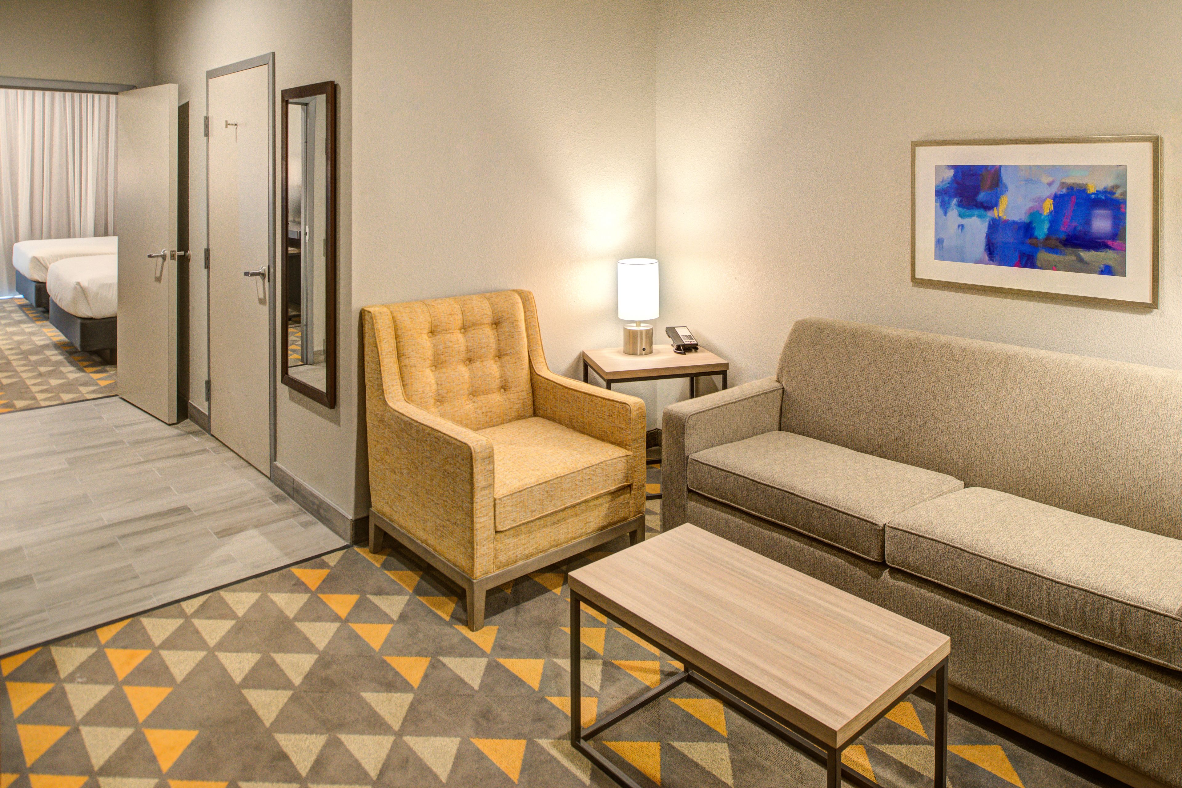 A hotel room with a sofa, armchair, and a small table. There is a lamp on a side table and a painting on the wall.