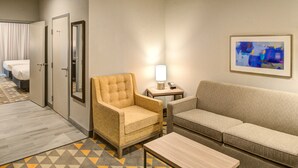 A hotel room with a sofa, armchair, and a small table. There is a lamp on a side table and a painting on the wall.