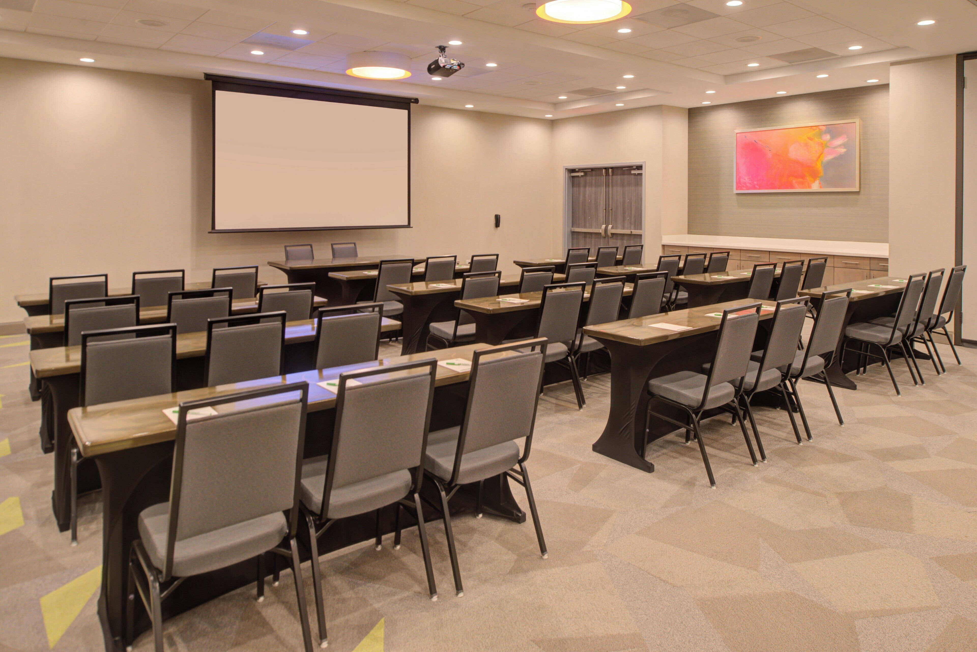 A conference room with multiple tables and chairs, a projector screen, and a large presentation screen.