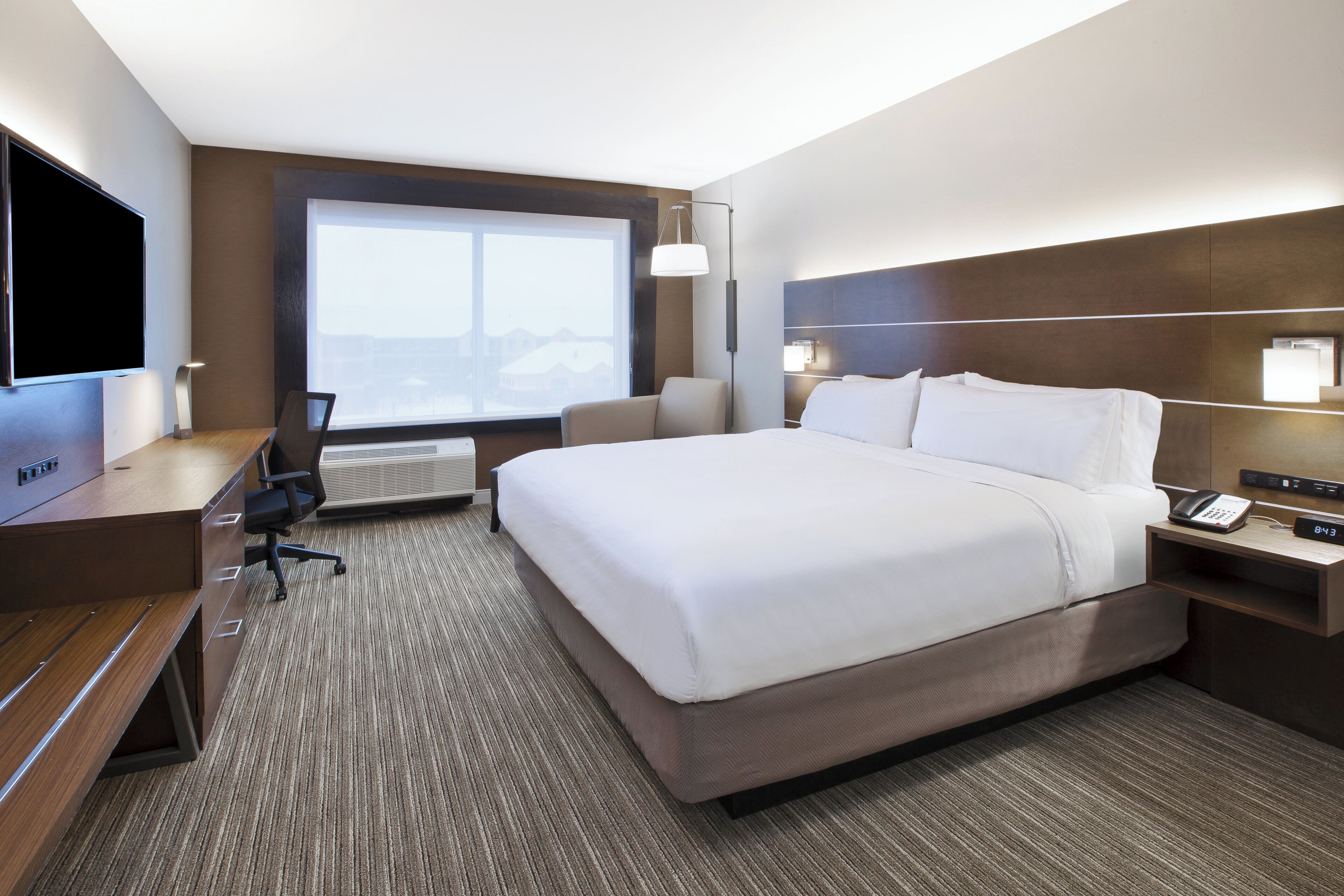 A hotel room with a large bed, a desk, a chair, a TV, and a window with a view of buildings.