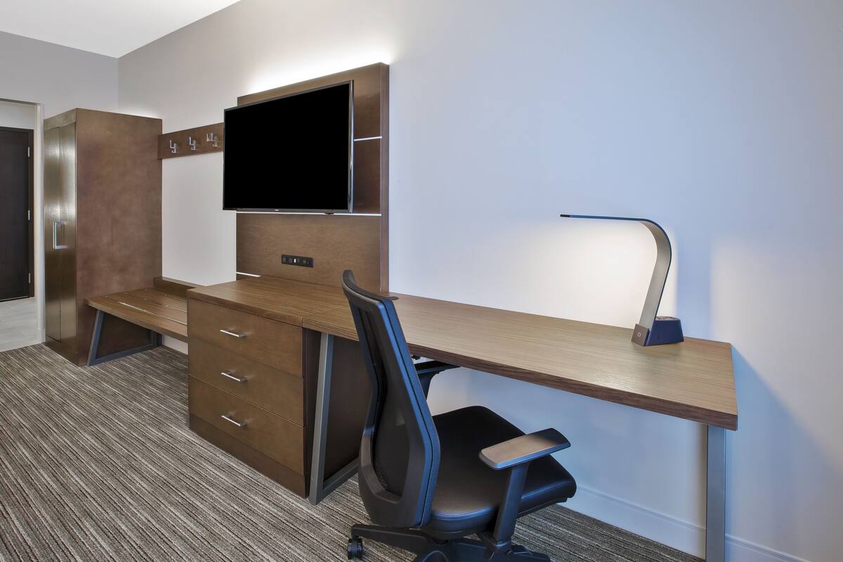 A modern hotel room with a wooden desk, a flat-screen TV, a desk lamp, and a black office chair.