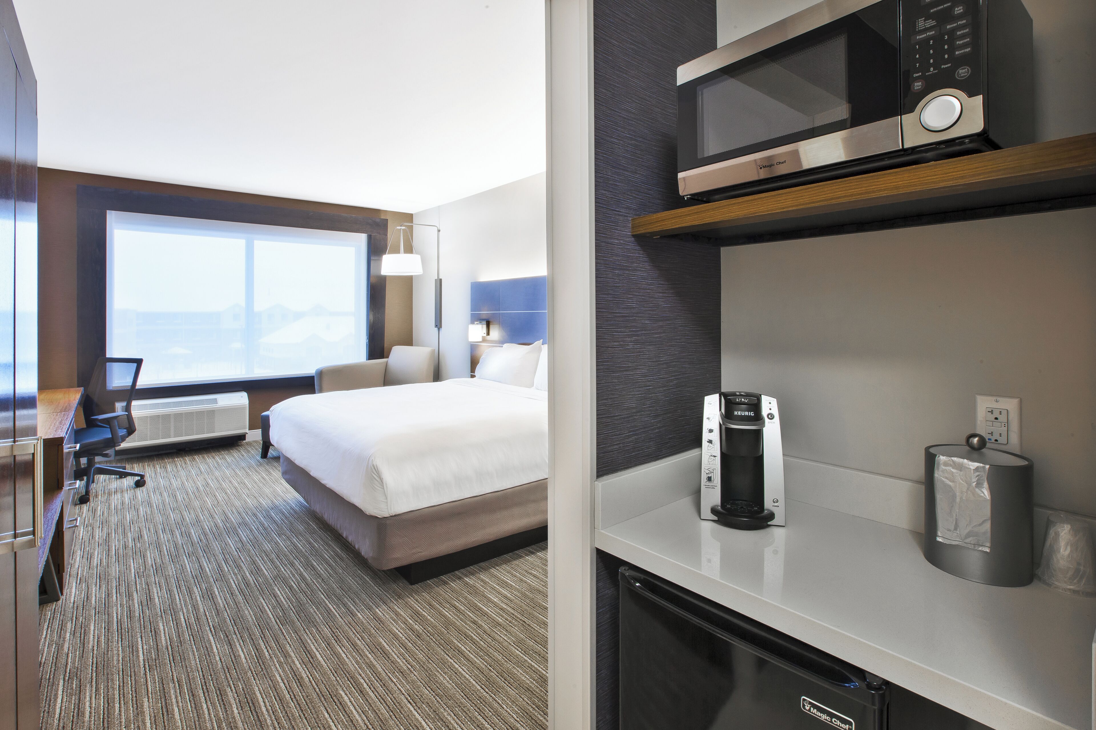 A hotel room with a bed, desk, chair, kitchenette, and a large window.