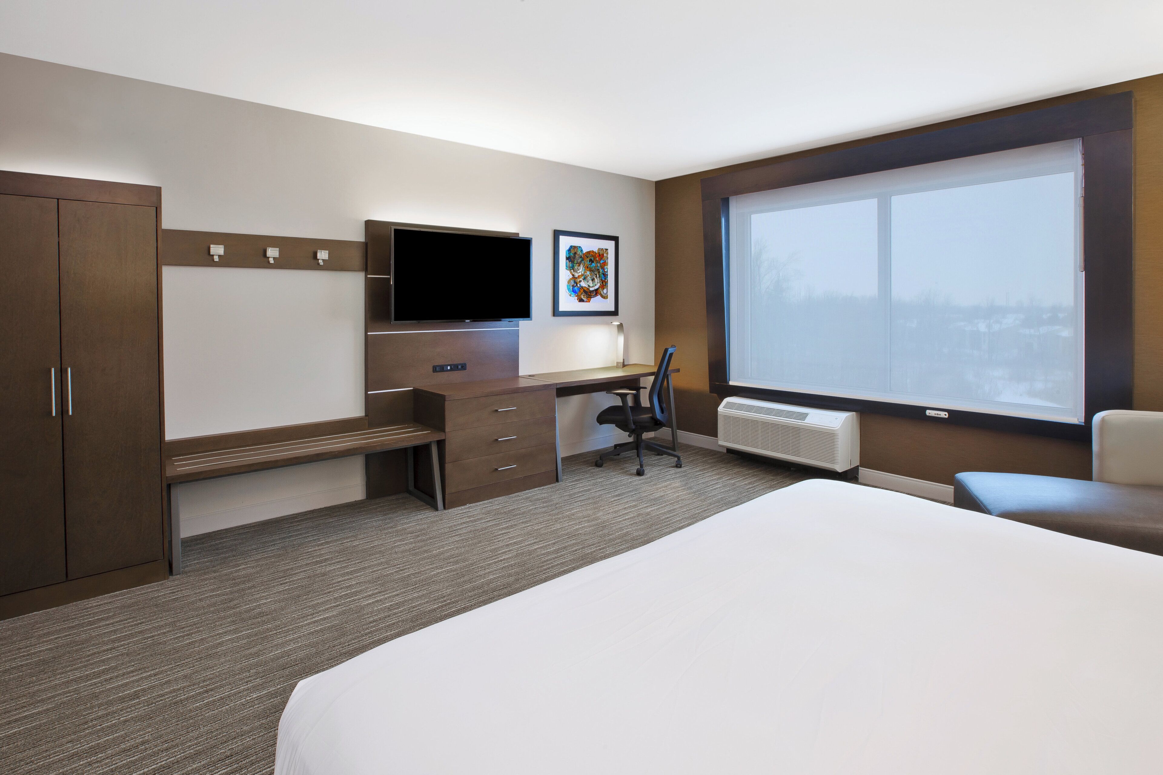 A hotel room with a bed, a desk with a chair, a television, a radiator, and a window.