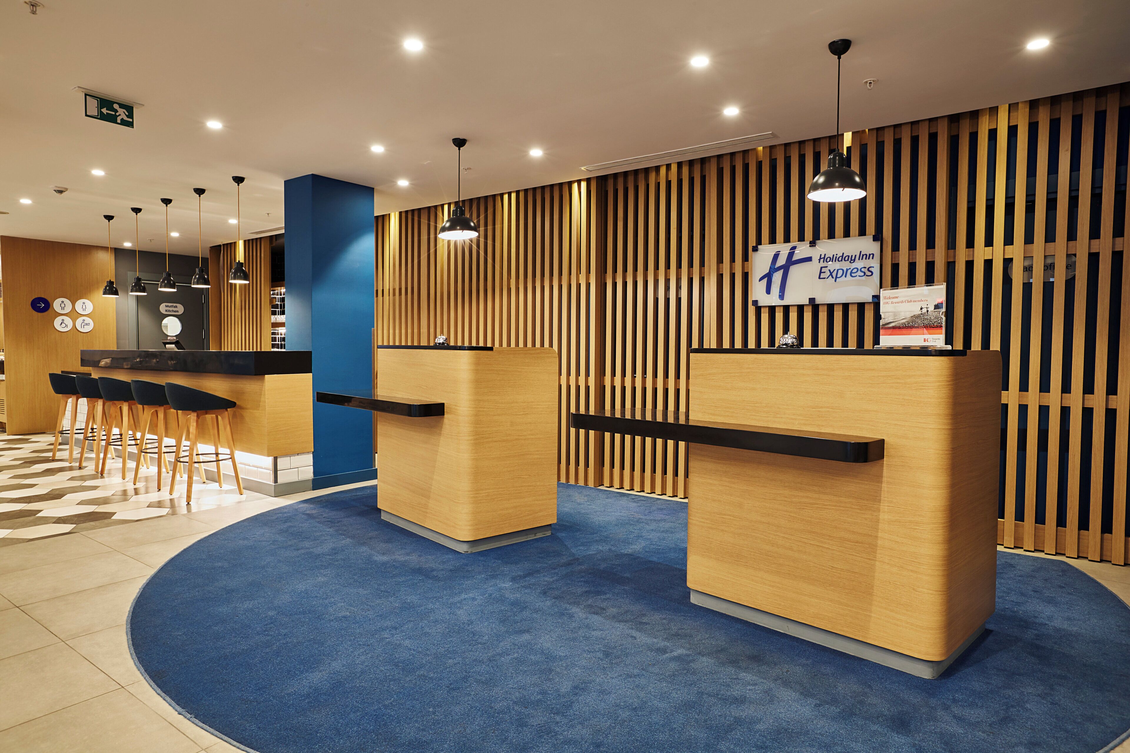 A hotel reception area with a blue carpet, wooden reception desks, and a vertical wooden panel wall.