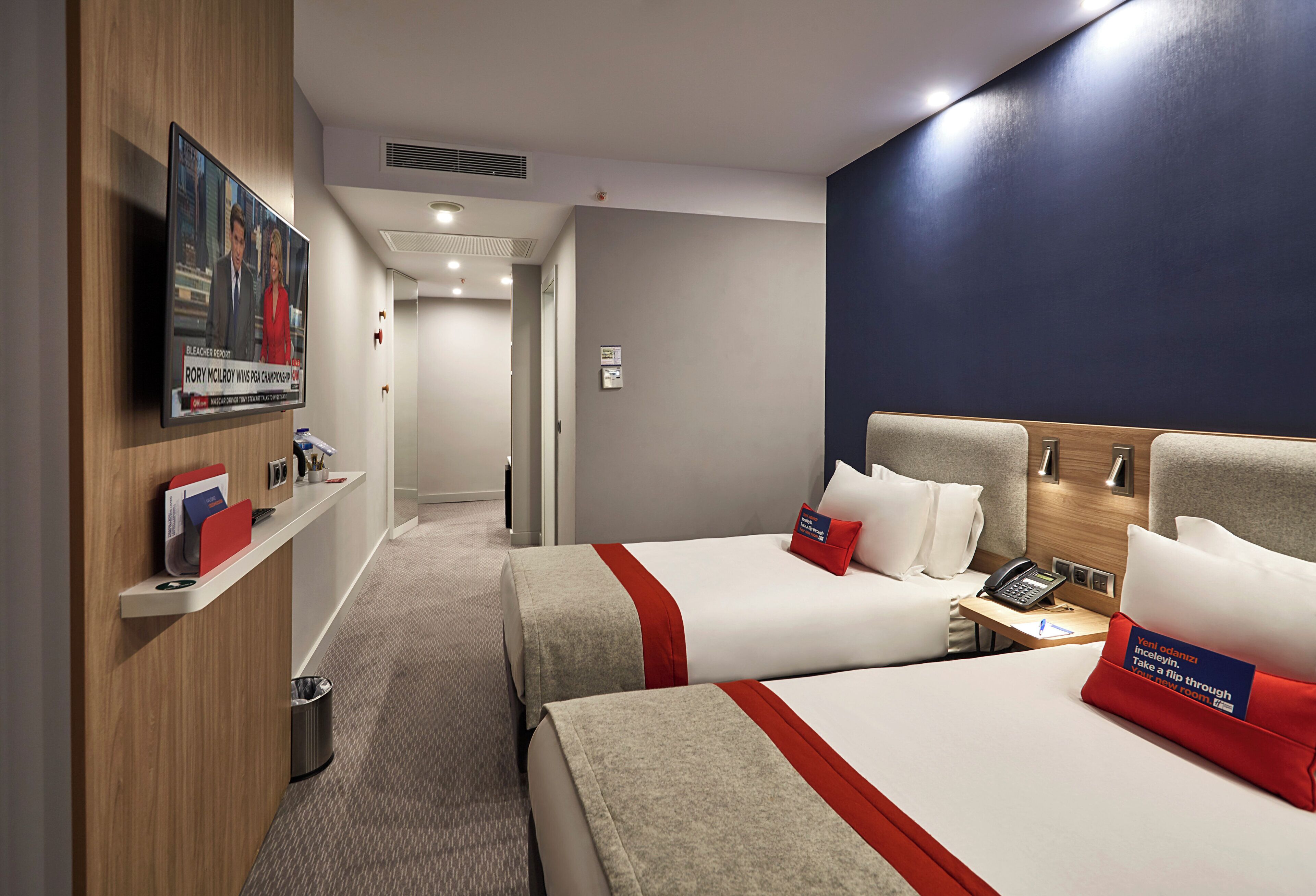 Ein Hotelzimmer mit zwei Betten, einem Schreibtisch mit Telefon, einem Wandfernseher und einem Mülleimer.