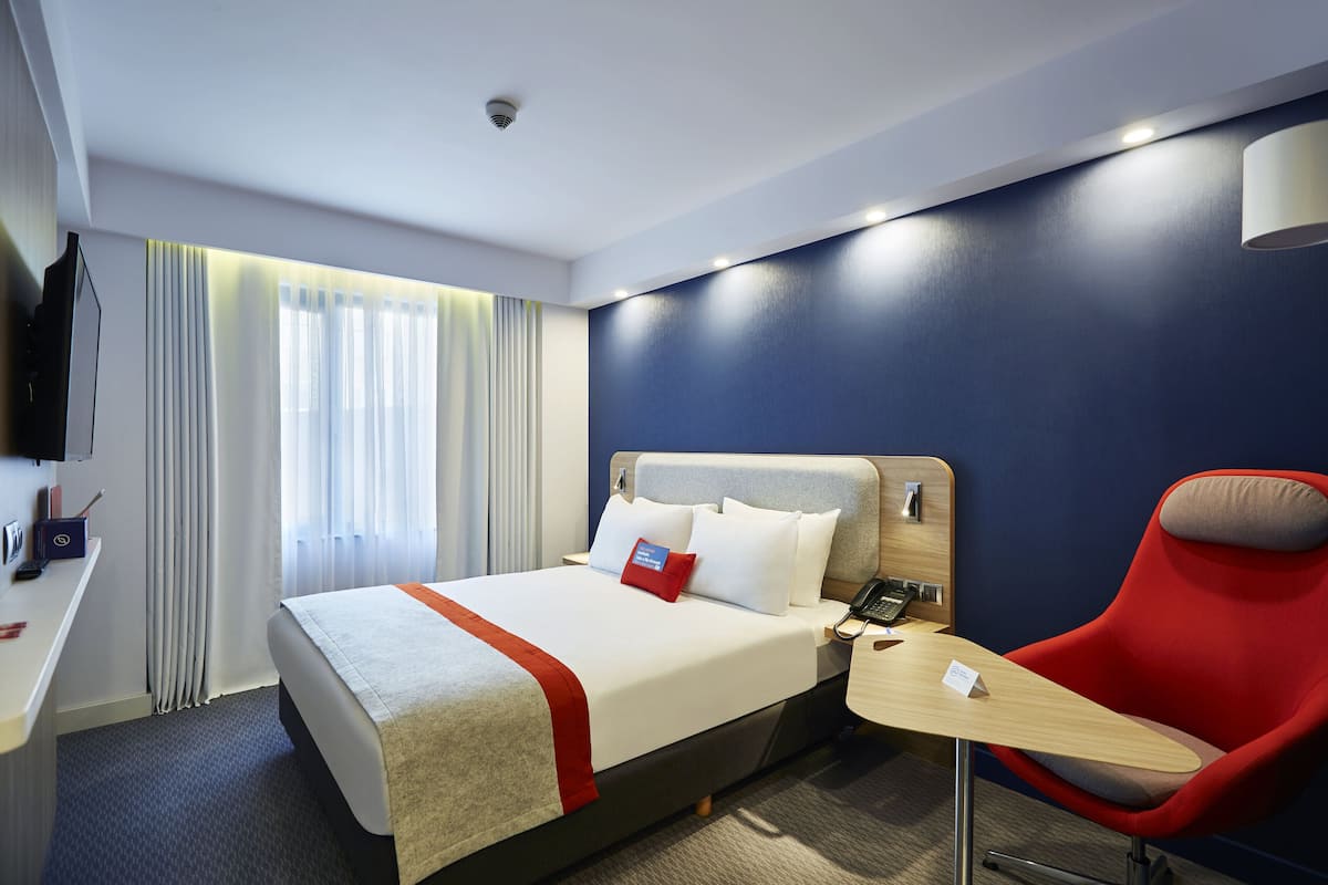 A hotel room with a bed, a desk, a chair, a TV, and a window with curtains.