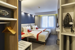 A hotel room with two beds, a desk, a mirror, a coat rack, and a security camera.