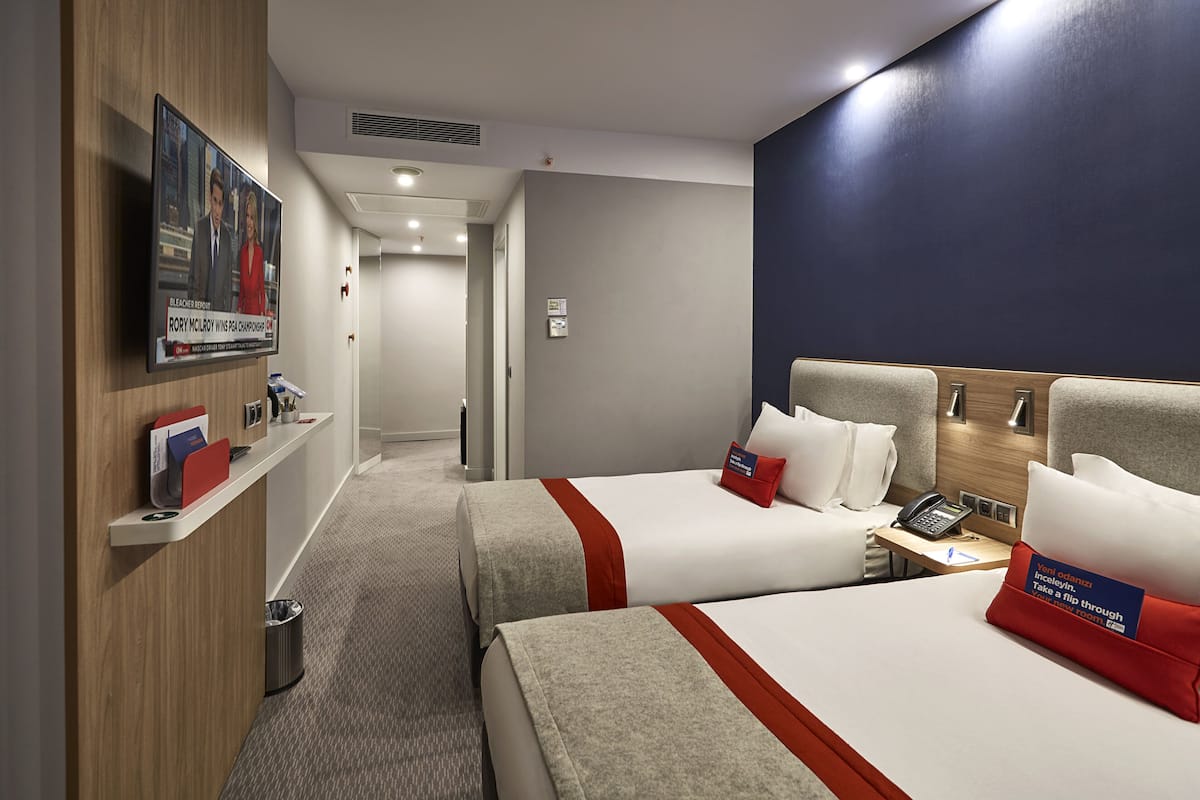 A hotel room with two beds, a TV mounted on the wall, a desk with a phone, and a trash can.