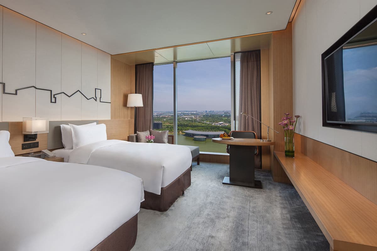 Een hotelkamer met twee bedden, een bureau, een stoel, een lamp, een vaas met bloemen en een groot raam met uitzicht op de stad.