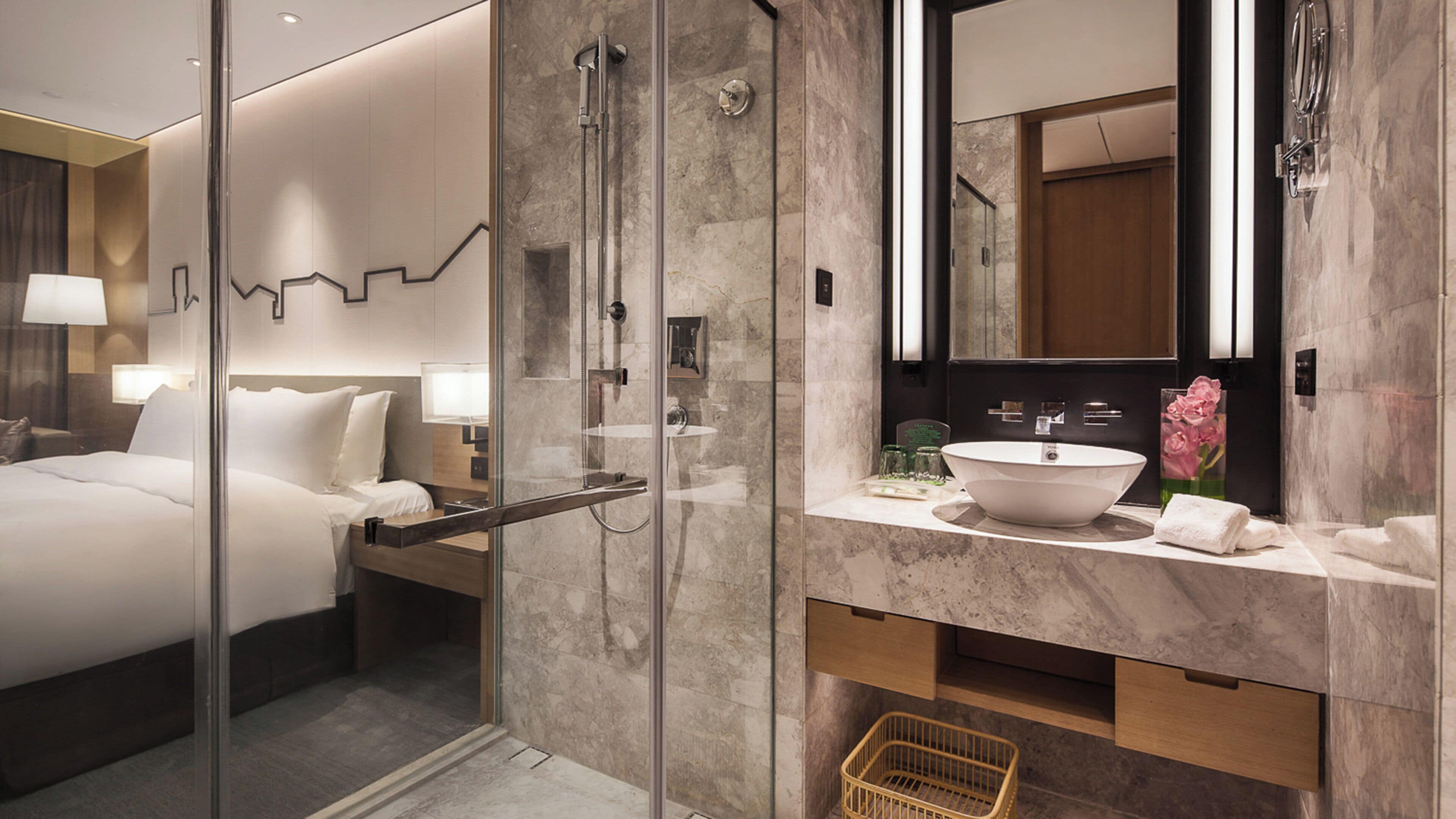 Un moderno baño de hotel con ducha de vidrio, un espejo grande, un lavamanos blanco y un tocador de madera.