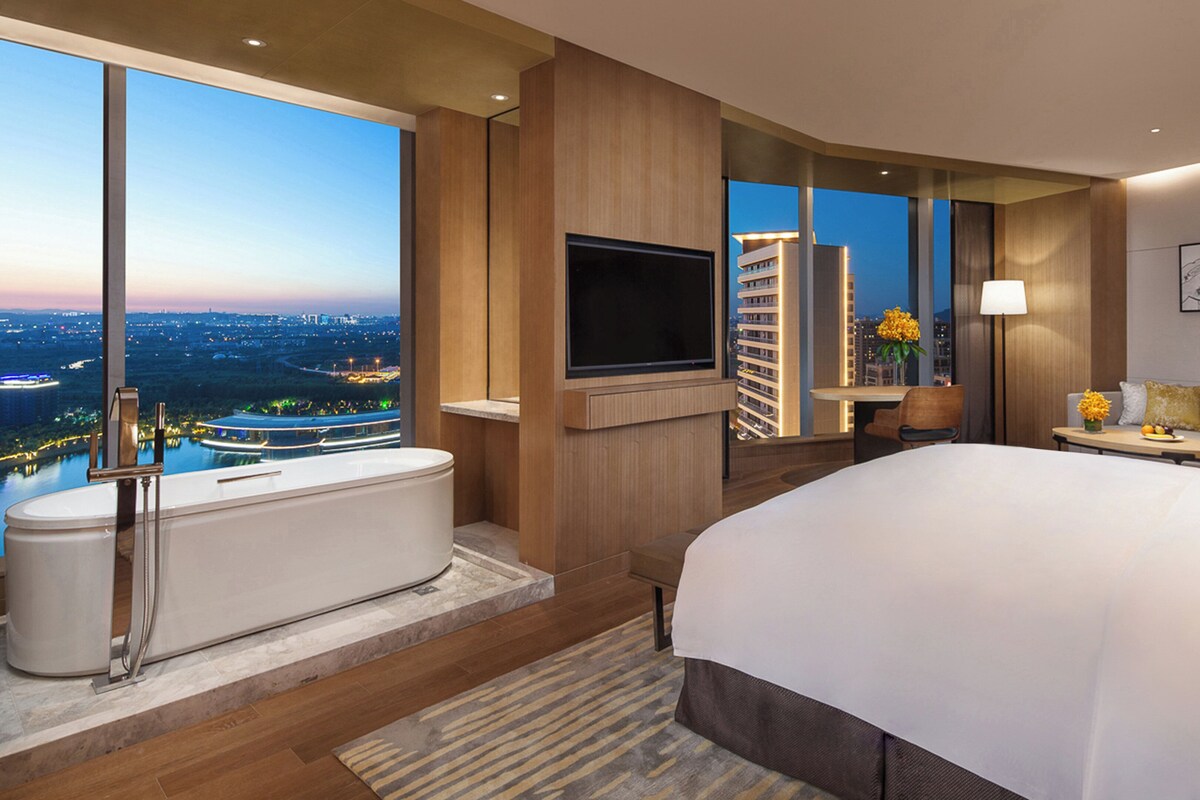Habitación de hotel moderna con una cama grande, bañera, televisor de pantalla plana y vista a la ciudad.