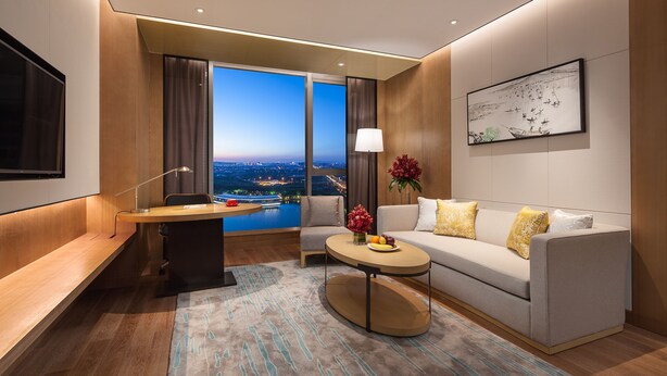 Una habitación de hotel moderna con un ventanal grande que ofrece vistas a la ciudad, un sofá con cojines decorativos, un escritorio con lámpara y una mesita con un jarrón de flores.
