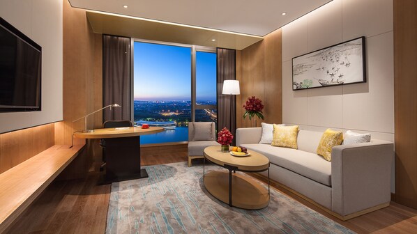 Una habitación de hotel moderna con un ventanal grande que ofrece vistas a la ciudad, un sofá con cojines decorativos, un escritorio con lámpara y una mesita con un jarrón de flores.