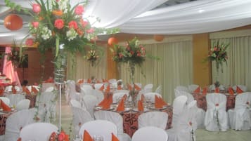 Salão de banquetes com mesas redondas montadas para um evento formal, decorado com toalhas brancas e guardanapos laranja.