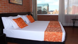 A hotel room with a large bed, white bedding, orange decorative pillows, and a view of the city through large windows. - Hotel Bogota Astral (Bogotá)